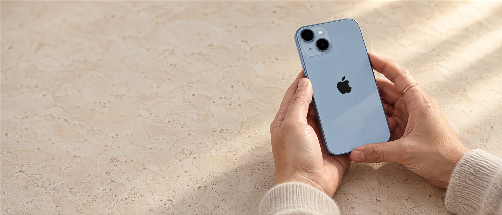 Editorial Revivo image: hands carefully holding a pale blue iPhone on warm travertine.